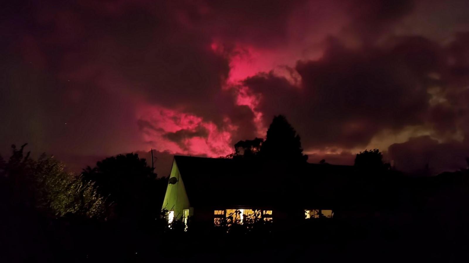 The northern lights in a rich red/pinkish colour, with clouds covering part of it above a large house and trees covered in shadow.