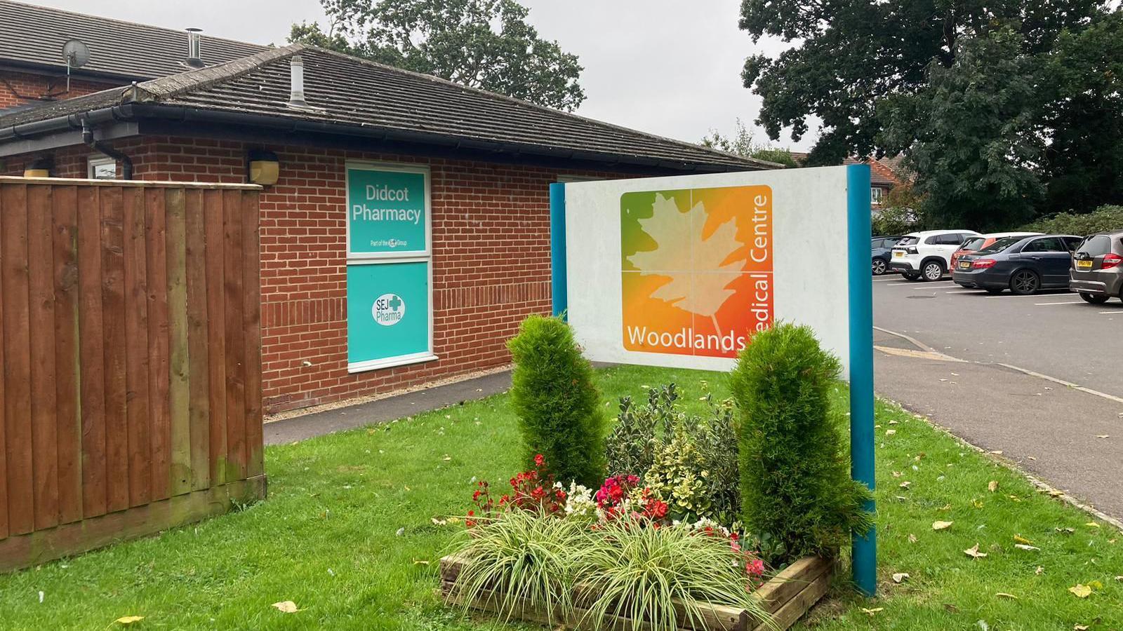 A sign saying 'Woodlands Medical Centre' with shrubbery around it, on a grass verge with a brick building and a car park in the background.