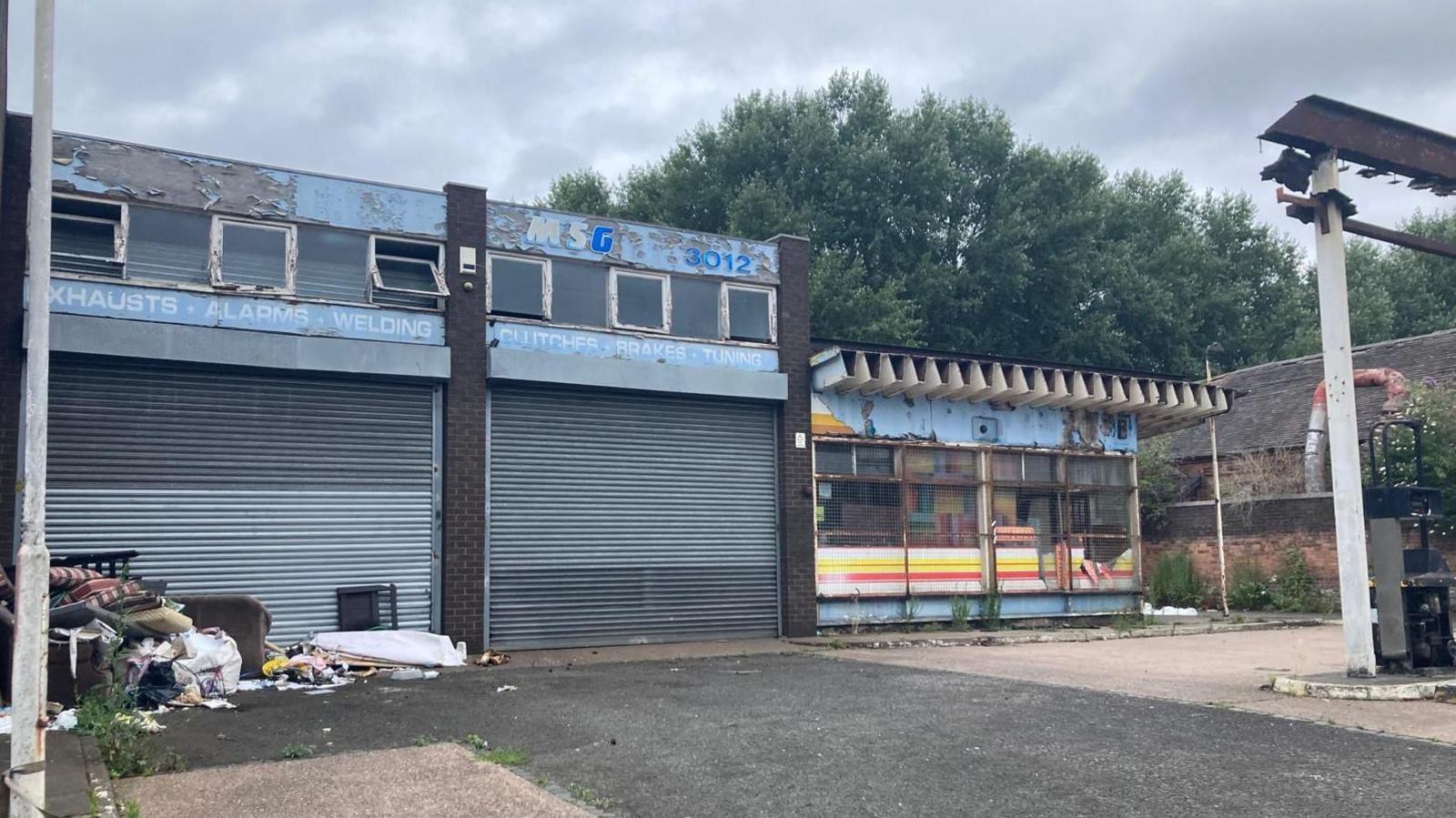 A derelict industrial building with metal shutters and rubbish scattered outside. 