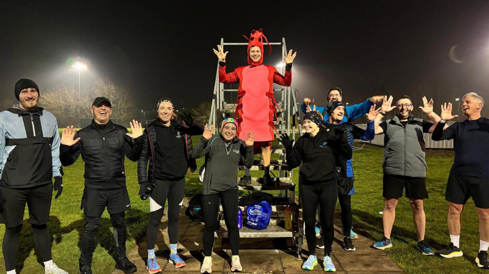 Jamie Campbell is standing in the middle of the picture, and is on some stairs. There are nine of his running friends standing around him with their hands in the air. 