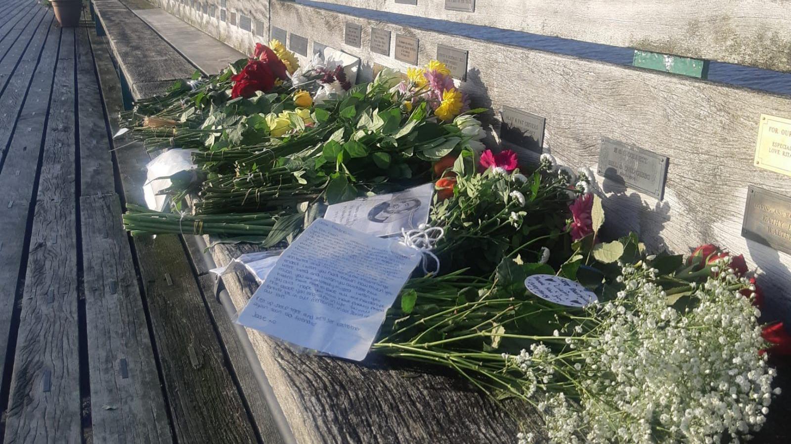 Several bouquets of flowers, letters and cards left in memory of Liam Payne. They have been placed on a bench on Clevedon Pier. Several plaques can be seen on the bench. 