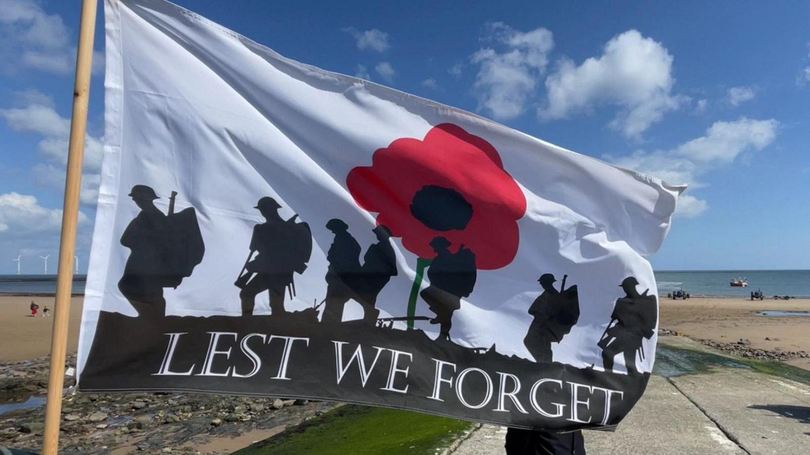 A flag saying 'lest we forget' flying in the forefront, with a beach behind and an man in military garb in a wheelchair