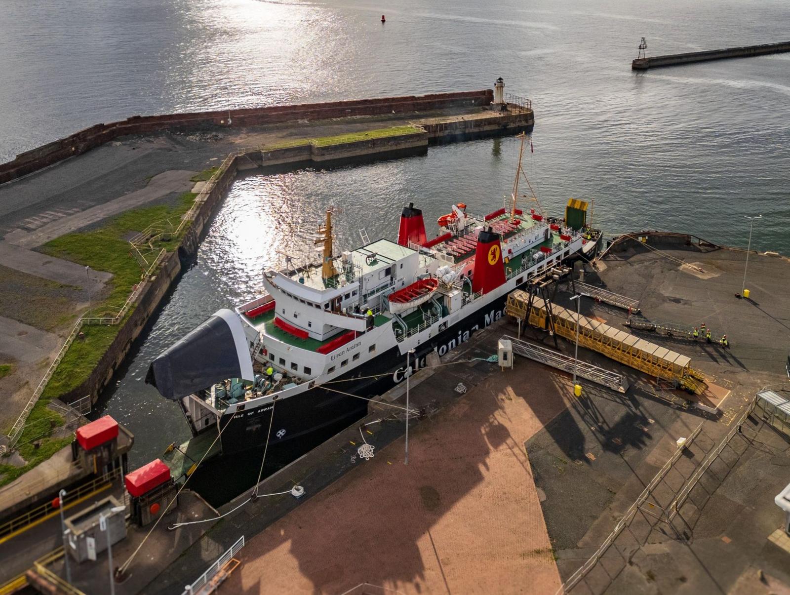 A black and white ship with red funnels berthed at a harbour. The harbour wall
