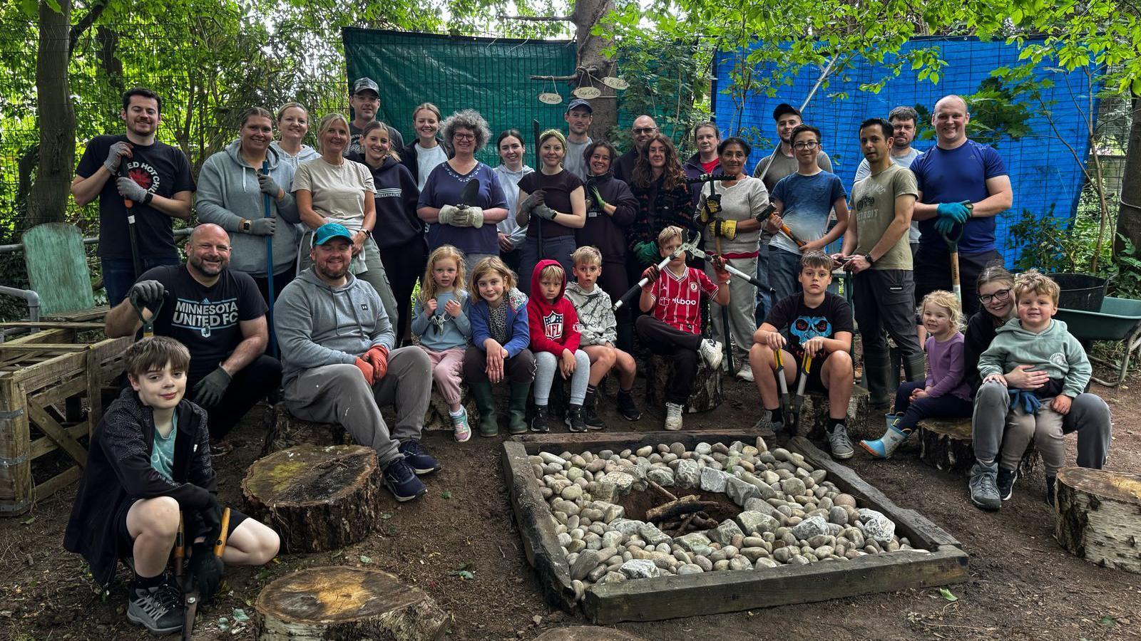 A group of adults and children are sitting around a campfire space. Some of them are holding gardening tools or wearing gloves.