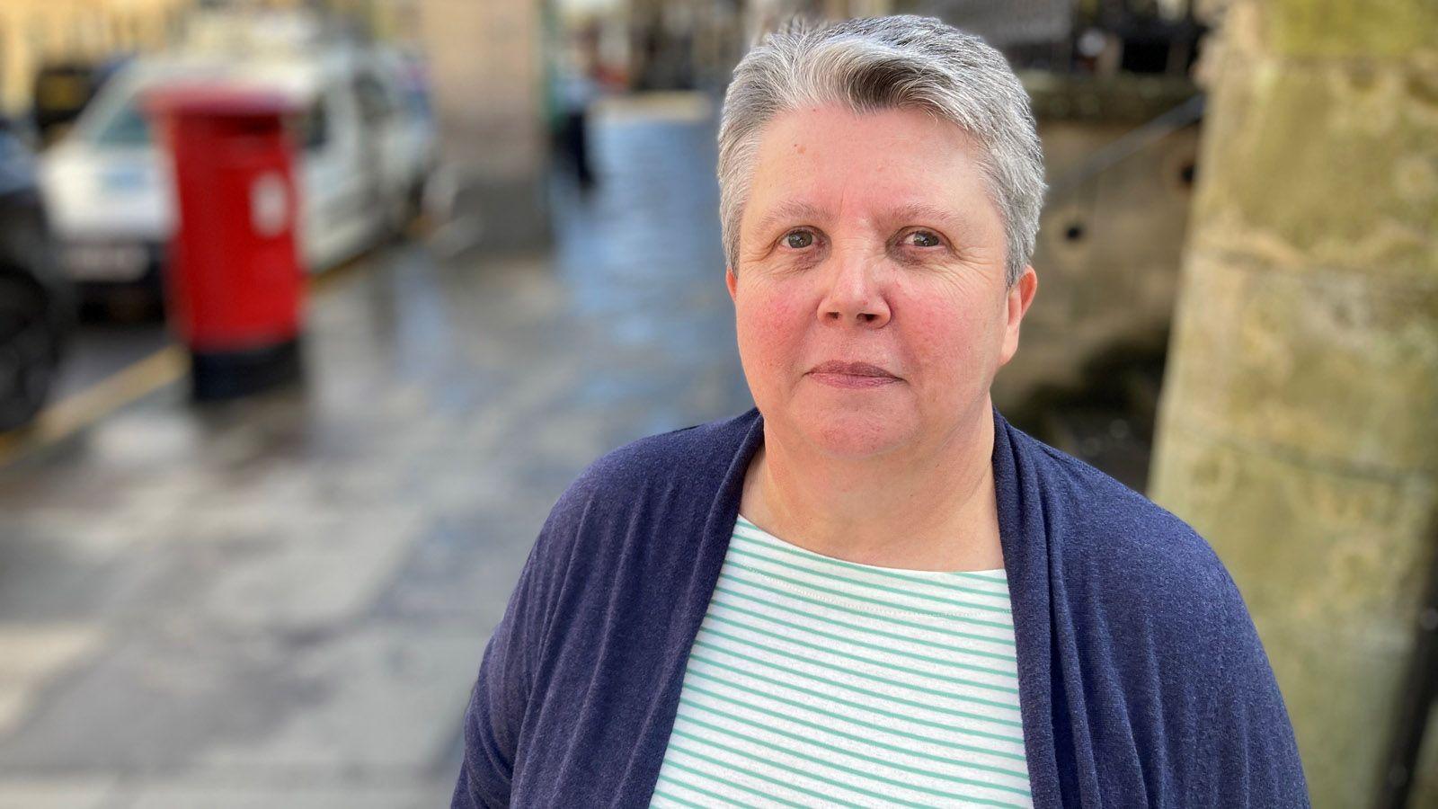 Fiona McClay has short, grey hair and is wearing a purple cardigan over whit top with thin green horizontal stripes. She is standing in a street and looking at the camera.