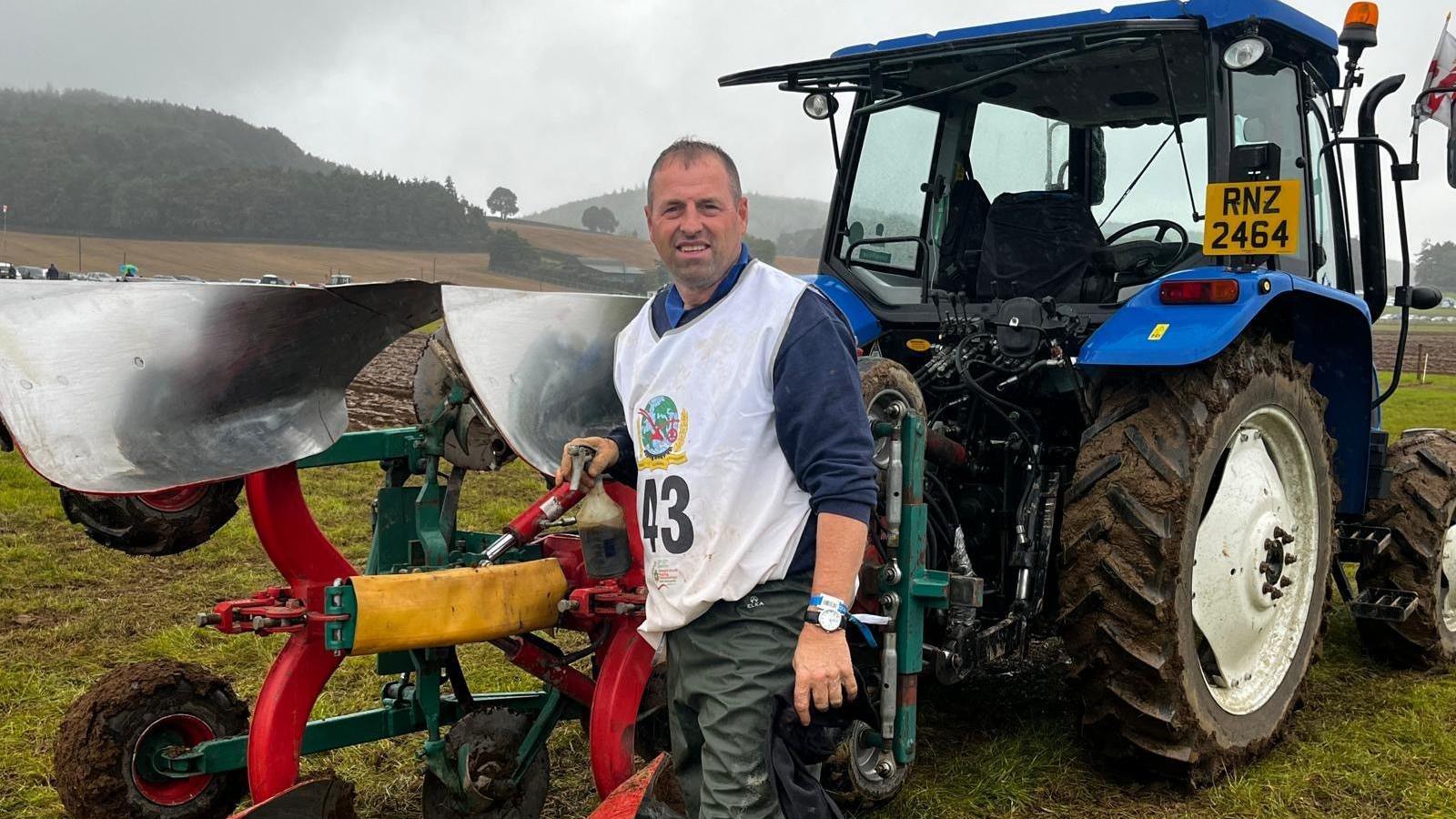 A man stands in front of a blue tractor with plouging equipment attached. He is in a field, with grey skies and hills behind. 