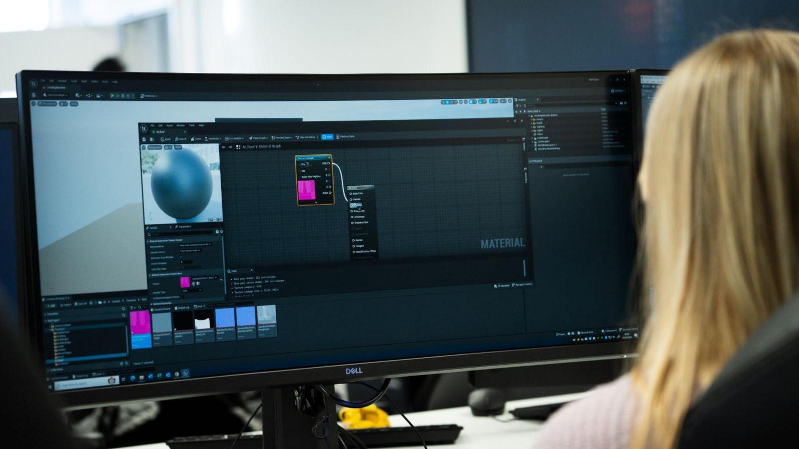 Part of the rear of a woman's head and shoulders, as she sits in front of computer screens. She has long, straight, fair hair. There are various programs open on the screen.