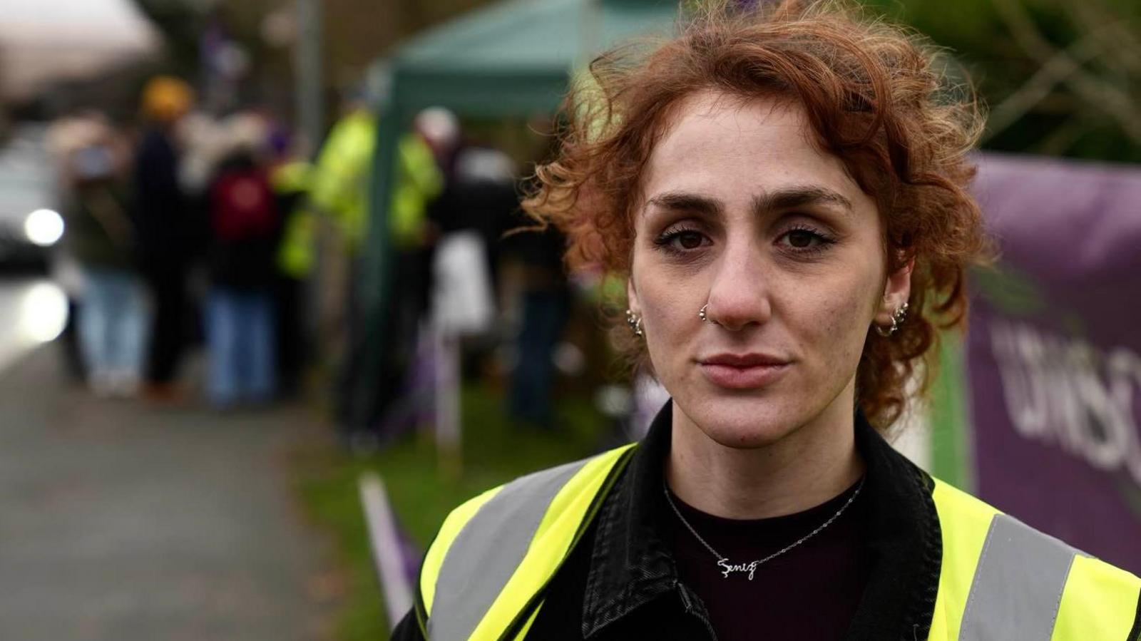 Shen Batmaz with auburn hair wears a hi-vis vest and stands on a picket line. She is looking straight at the camera with a serious expression. In the background a number of people are standing by a road. 