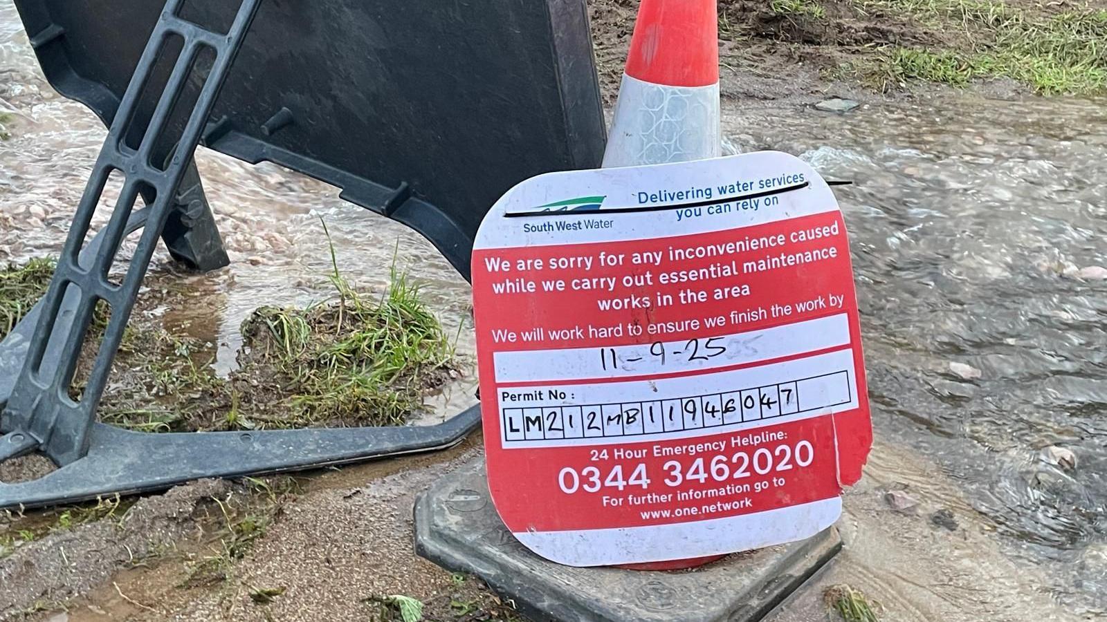 A traffic cone with a red and white South West Water sign attached to it. It contains information on when the work should be finished - 11.09.25. Behind the sign is the back of another sign and flowing water along the ground. 