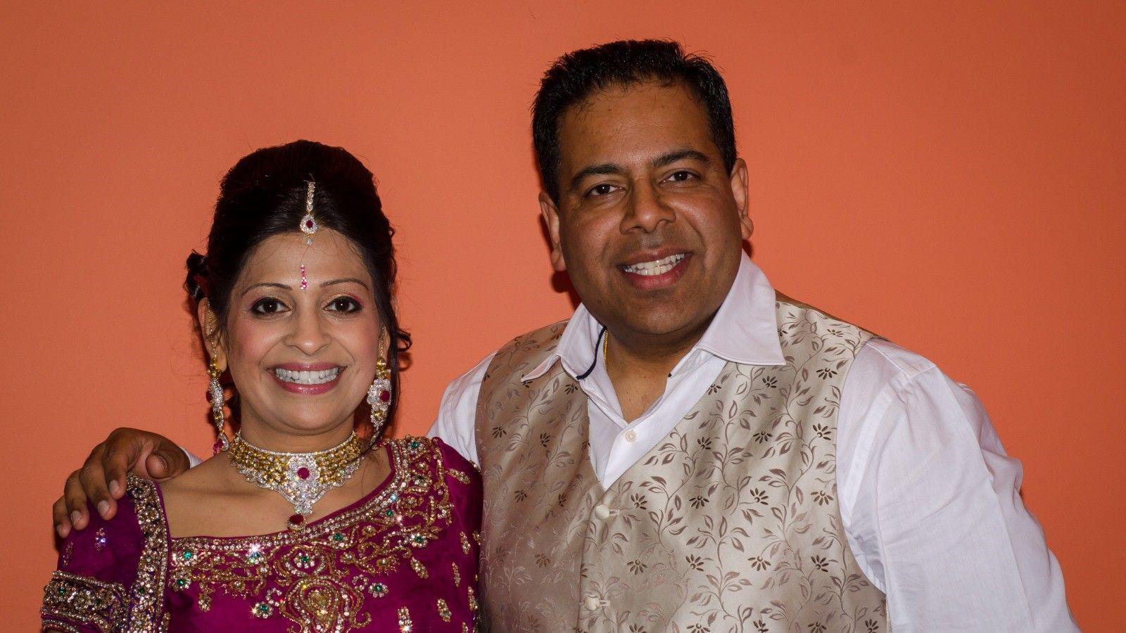 A photograph of Leela - on the left - and Manoj Keshavji with an orange background behind them. Leela is wearing a red-purple coloured dress with many jewels on and Manoj is wearing a white shirt and light gold coloured waistcoat with a leaf pattern on.