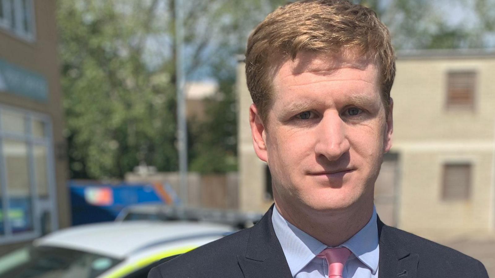 Kent Police and Crime Commissioner Matthew Scott stands in front of a police car.  He is a white man with strawberry blonde hair, wearing a suit and tie. There are buildings out of focus in the background. 