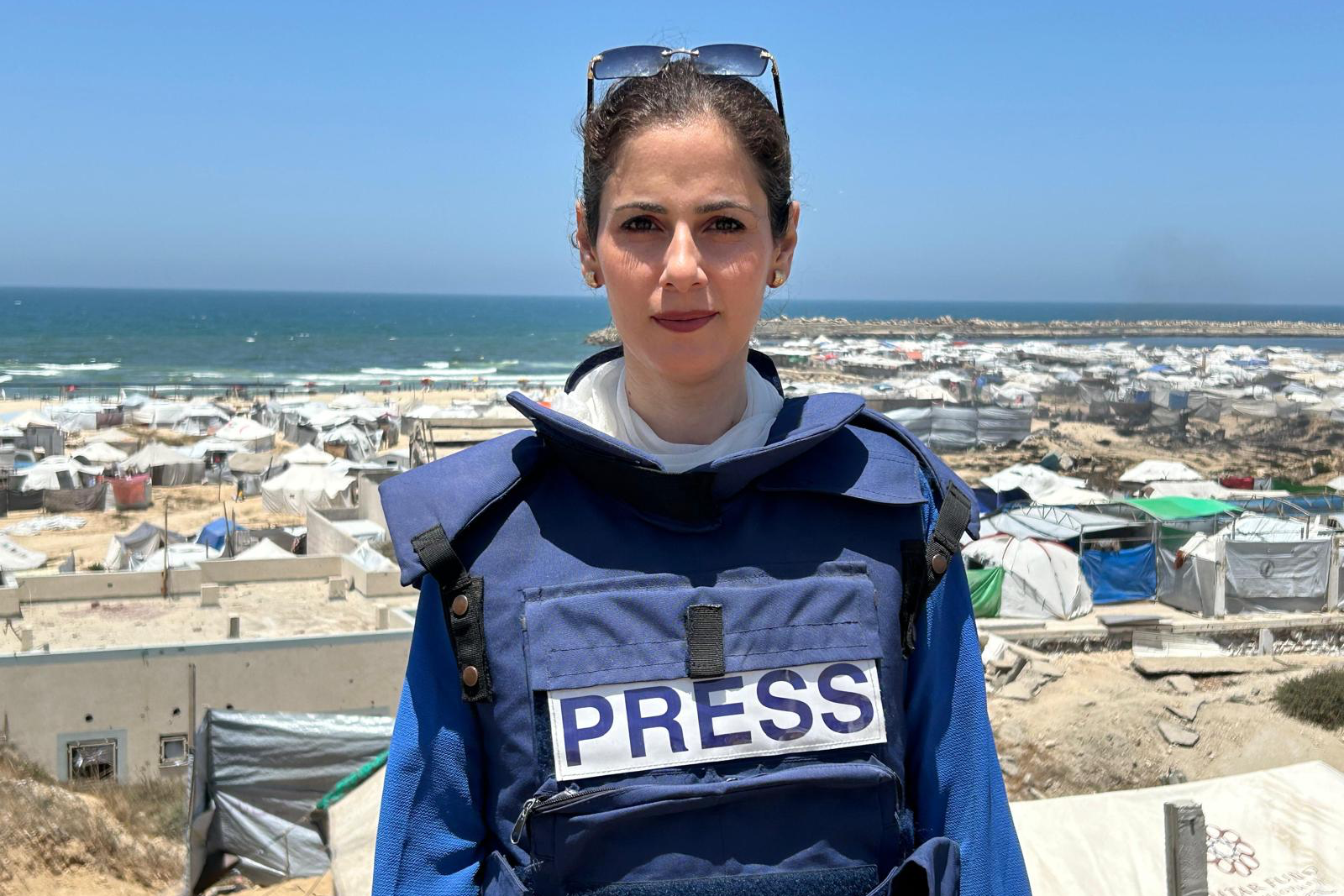 Ghada Al-Kurd stands in front of a mass of tents with the sea in the background. She is wearing a blue press flak jacket and has glasses on her head.