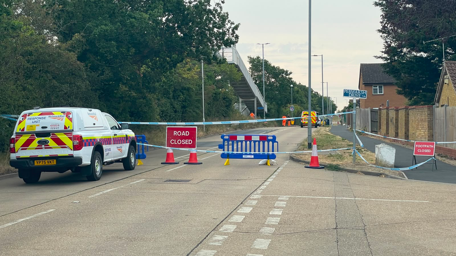 Part of a road closed off. Blue and white striped tape acts as a cordon and there are road and footway closure signs propped up on the ground. A Network Rail response unit van is parked outside of the cordon.