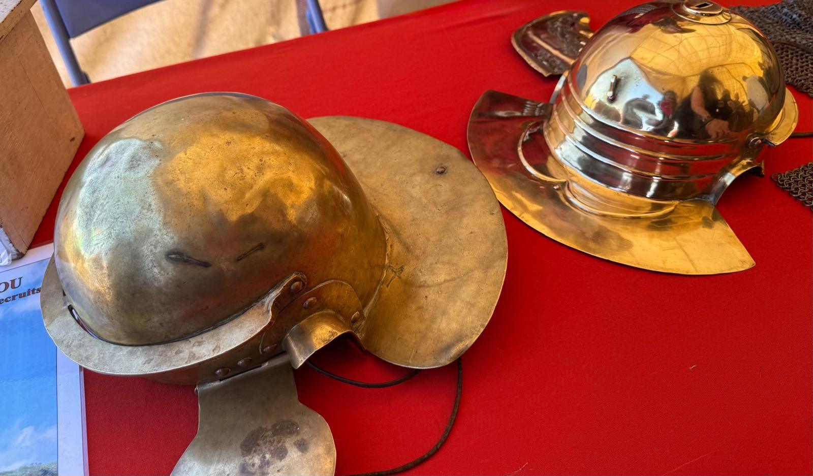 Two brass or gold helmets on a table inside a tent. They are placed on display, and one is much shinier than the other. 