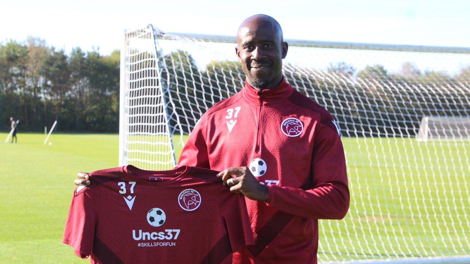 Adomah standing behind one of the goals at the Walsall training ground ewearing their training kit and holding a shirt that reads 'uncs37 #SKILLSFORFUN"