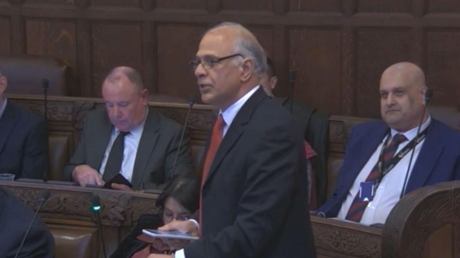 Pervez Ahktar stands in the wooden panelled council chamber speaking into a microphone surrounded by colleagues seated on wooden benched. He wears a black suit jacket, white shirt and red tie.