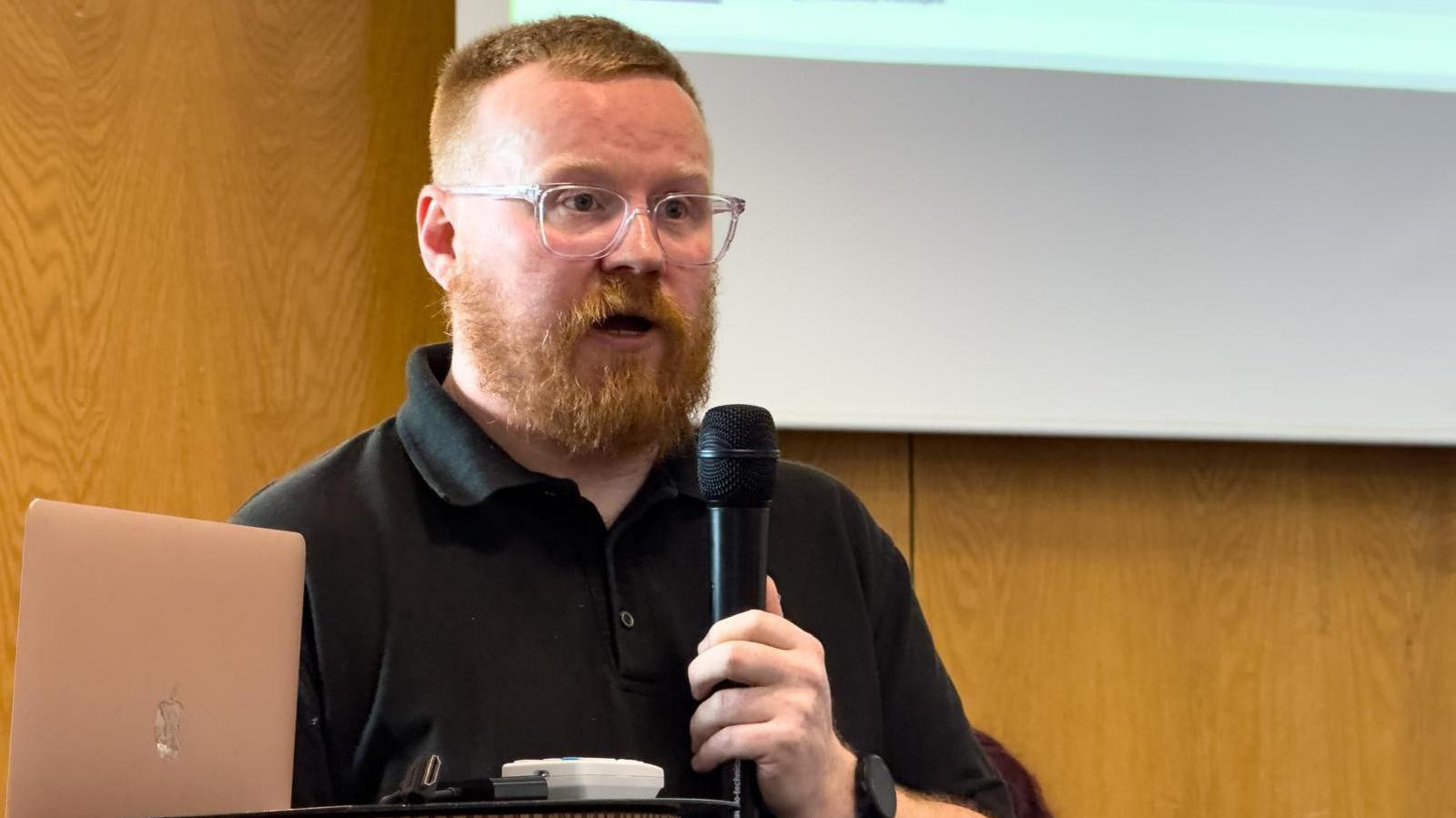 Rob Smith holding a microphone and speaking at the event. He has ginger hair and a beard and wears glasses and a black polo shirt. 