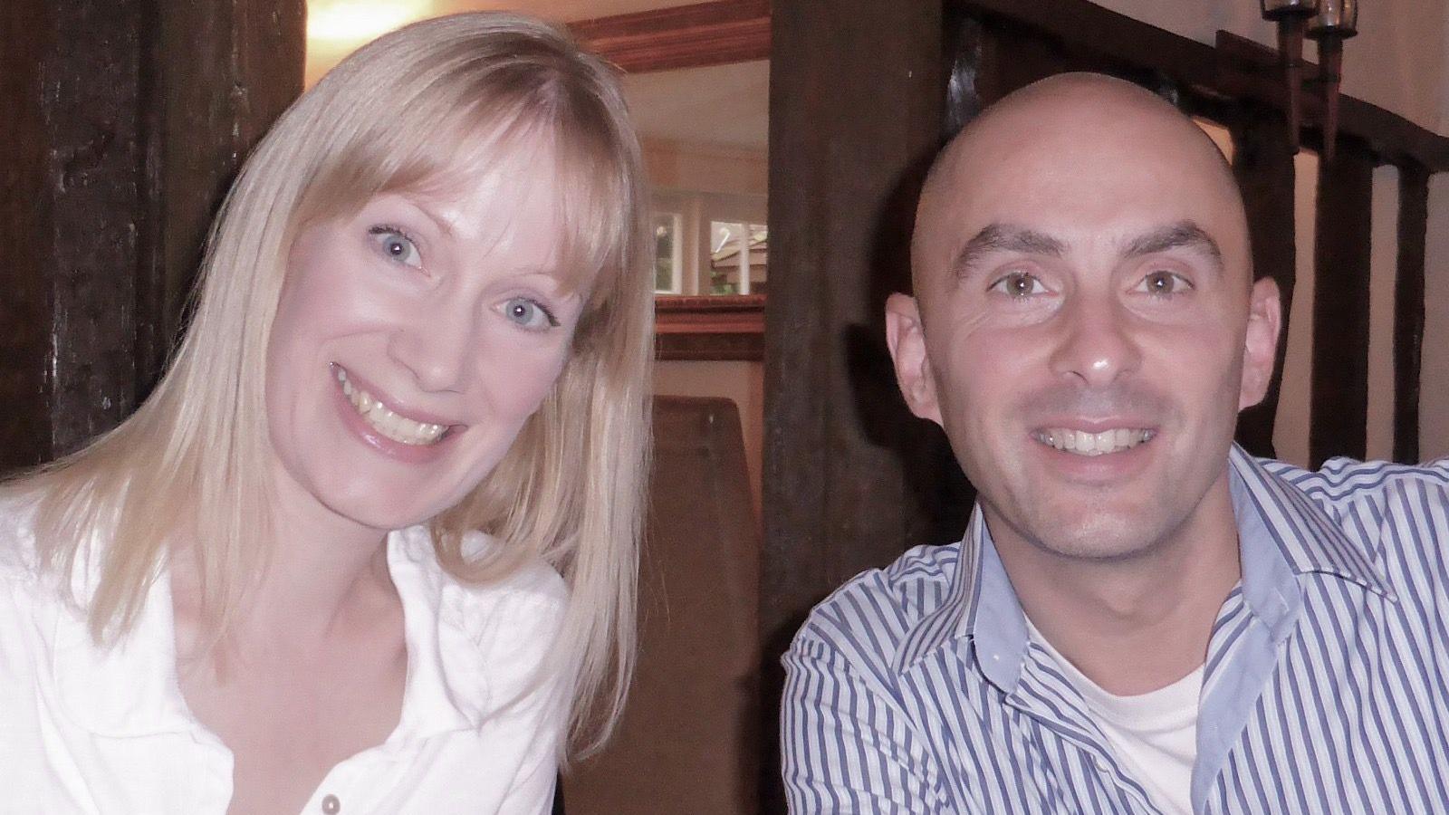 A woman in a white shirt and a bald man in a blue and white striped shirt. Both are looking at the camera and smiling.