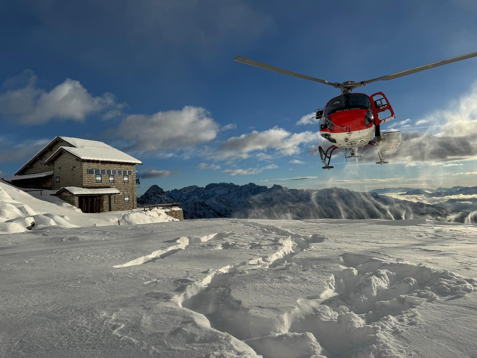 Red helicopter with white underbelly prepares to land on snow close to a lodge on a sunny day with blue sky and clouds