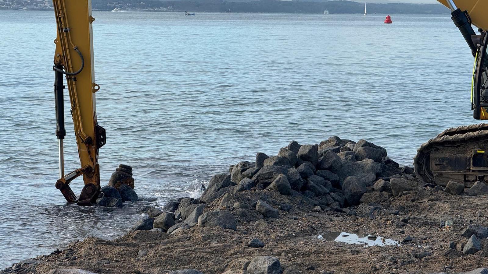 A yellow digger is lifting the ordnance out of the water along Southsea seafront, the shore is covered with rocks and there is the sea in the background