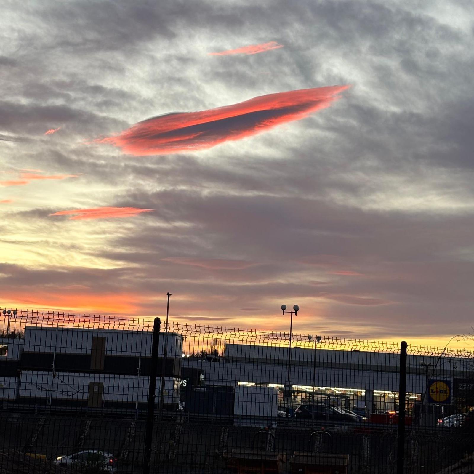 The cloud glows an orange-pink above industrial buildings that are behind a tall wire fence.