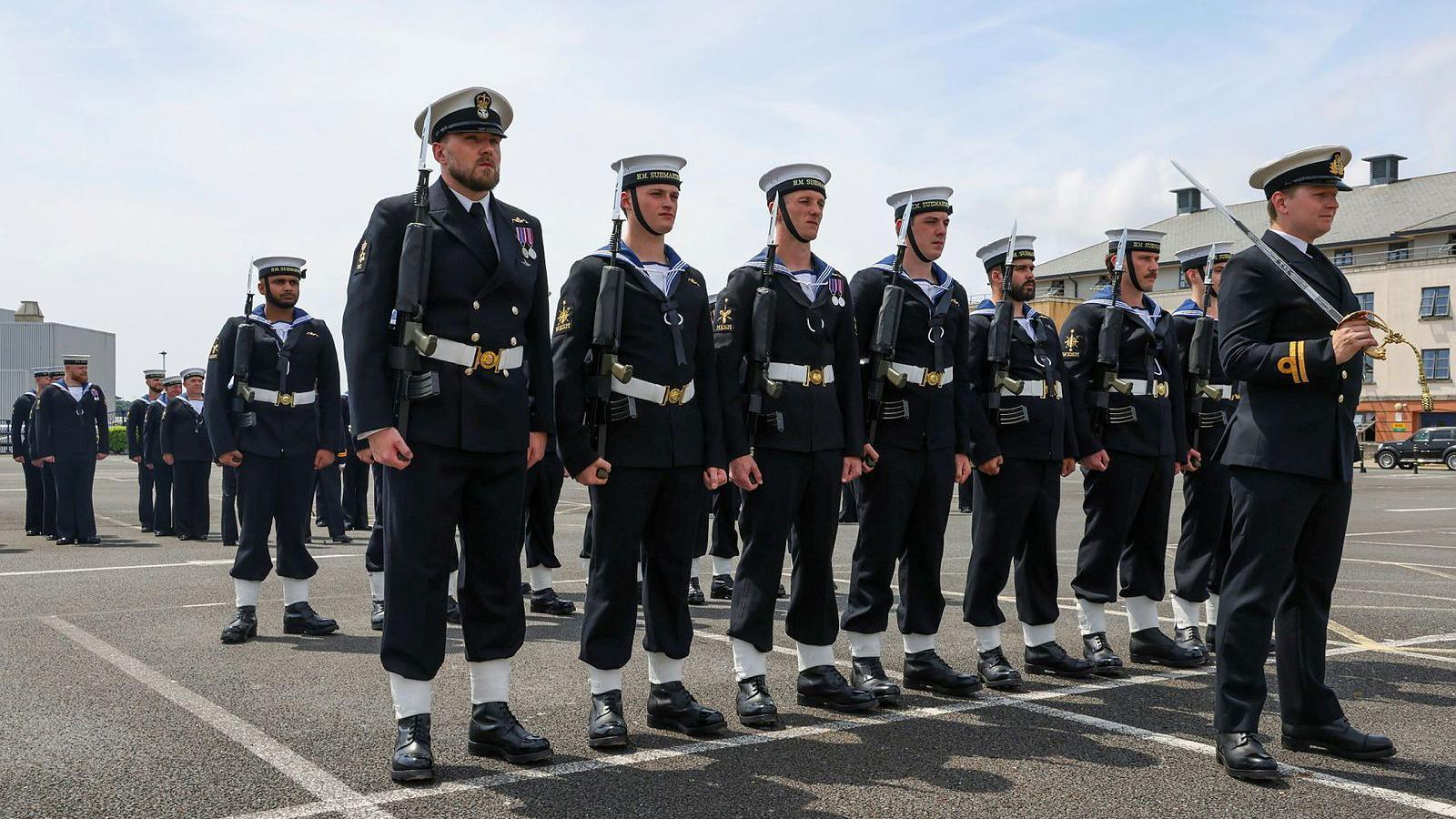 Royal Navy officers in uniform. There are srood in formation. The sky is blue with white clouds.