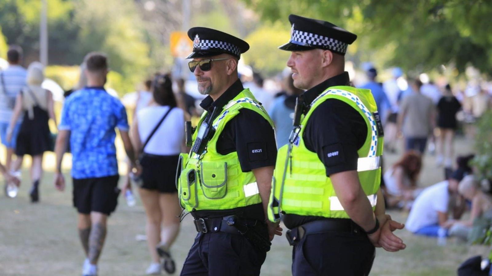 Two police officers, one wearing sun glasses, watch as Oasis fans walk by at Heaton Park