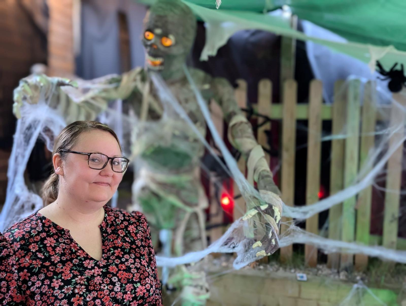 A woman stands in front of a model of a bandaged mummy. The woman has blonde hair and glasses, and she is wearing a black and red floral shirt. The mummy is decorated with fake cobwebs and has glowing yellow eyes.