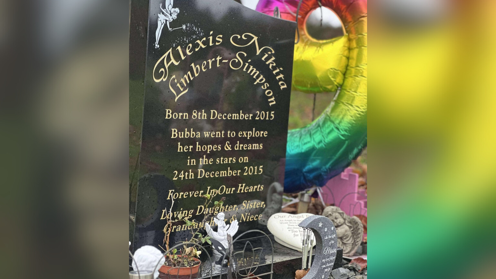 A black headstone in a cemetery, surrounded with ornaments and a rainbow-coloured balloon.