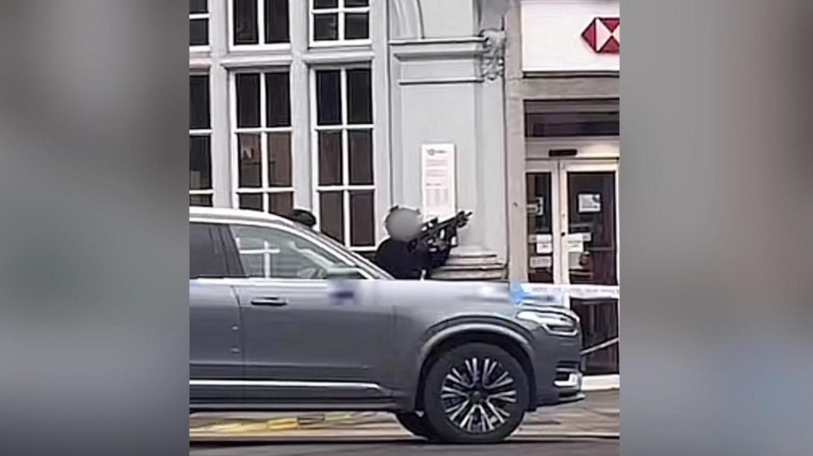 An armed police officer crouching down behind a silver car outside a branch of HSBC bank, aiming a gun upwards