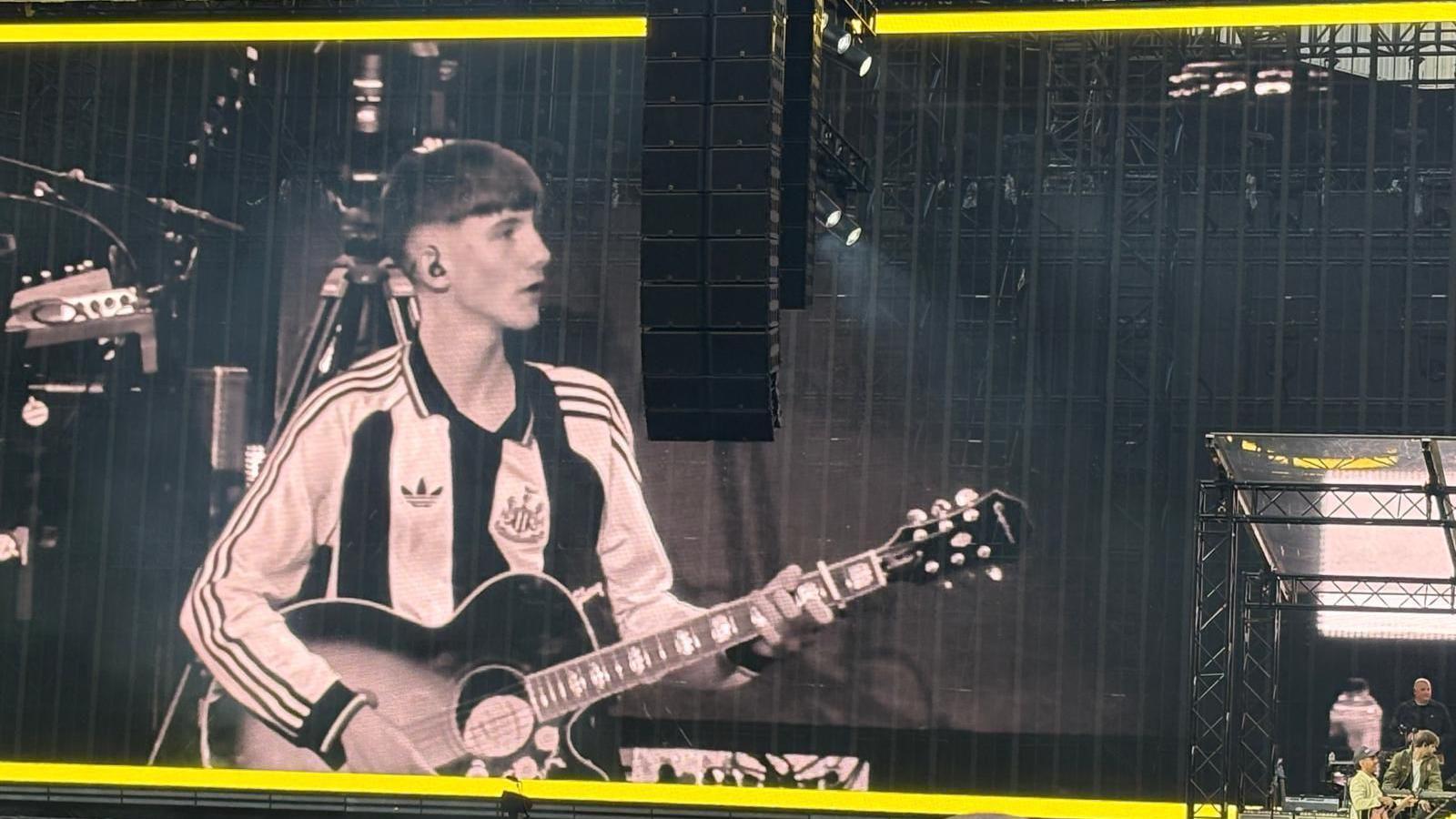 A picture of one of the screens inside St James' Park. The screen shows a close up of Joe wearing a Newcastle shirt and playing guitar on stage.
