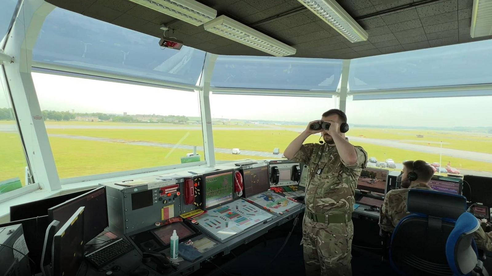 The visual control room at RAF Brize Norton. One man is standing and looking through a set of binoculars. Another man sits in front of a computer and looks at control panels, facing away from the camera. The green air field can be seen though the windows. 