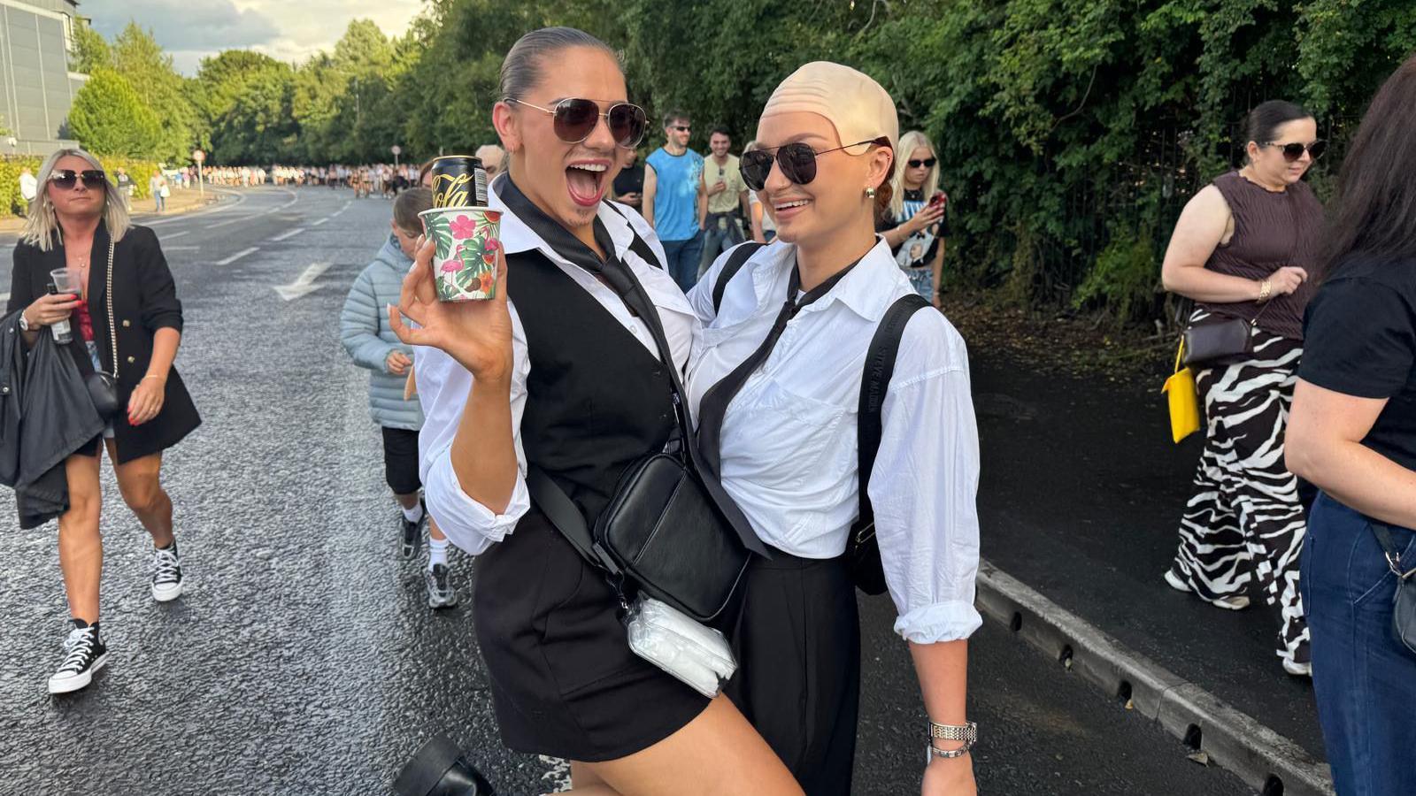 Two women. Both are wearing white shirts, black ties and sunglasses. The one on the right is wearing a bald cap and the one on the left is wearing a black waiscoat.