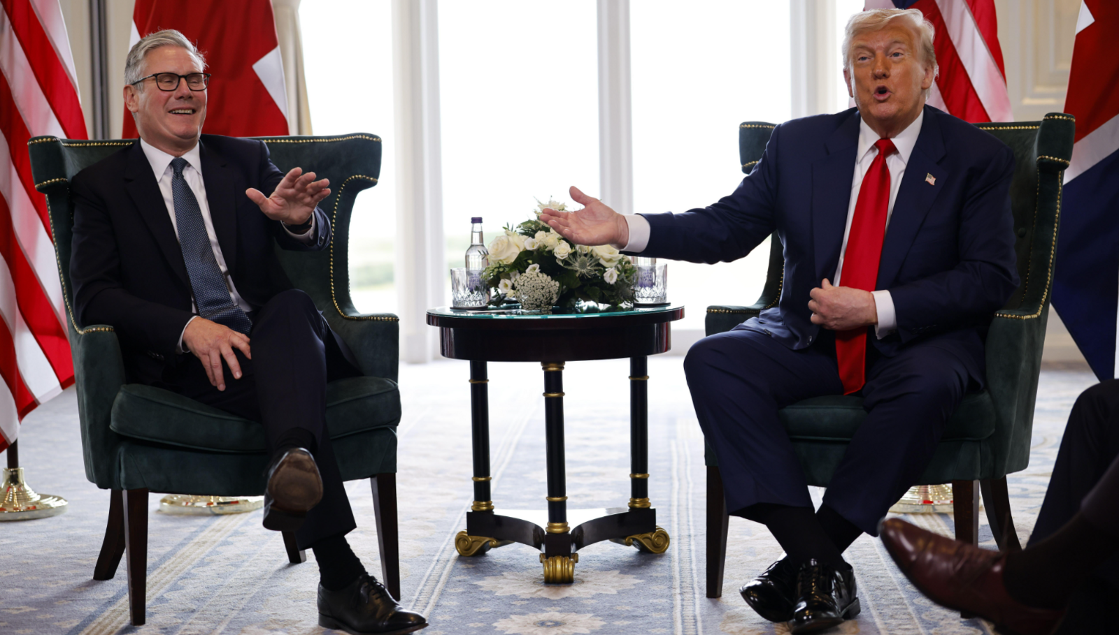 Trump and Starmer seated at the press conference. Starmer is smiling and Trump is gesturing towards him. There are US and UK flags in the background.