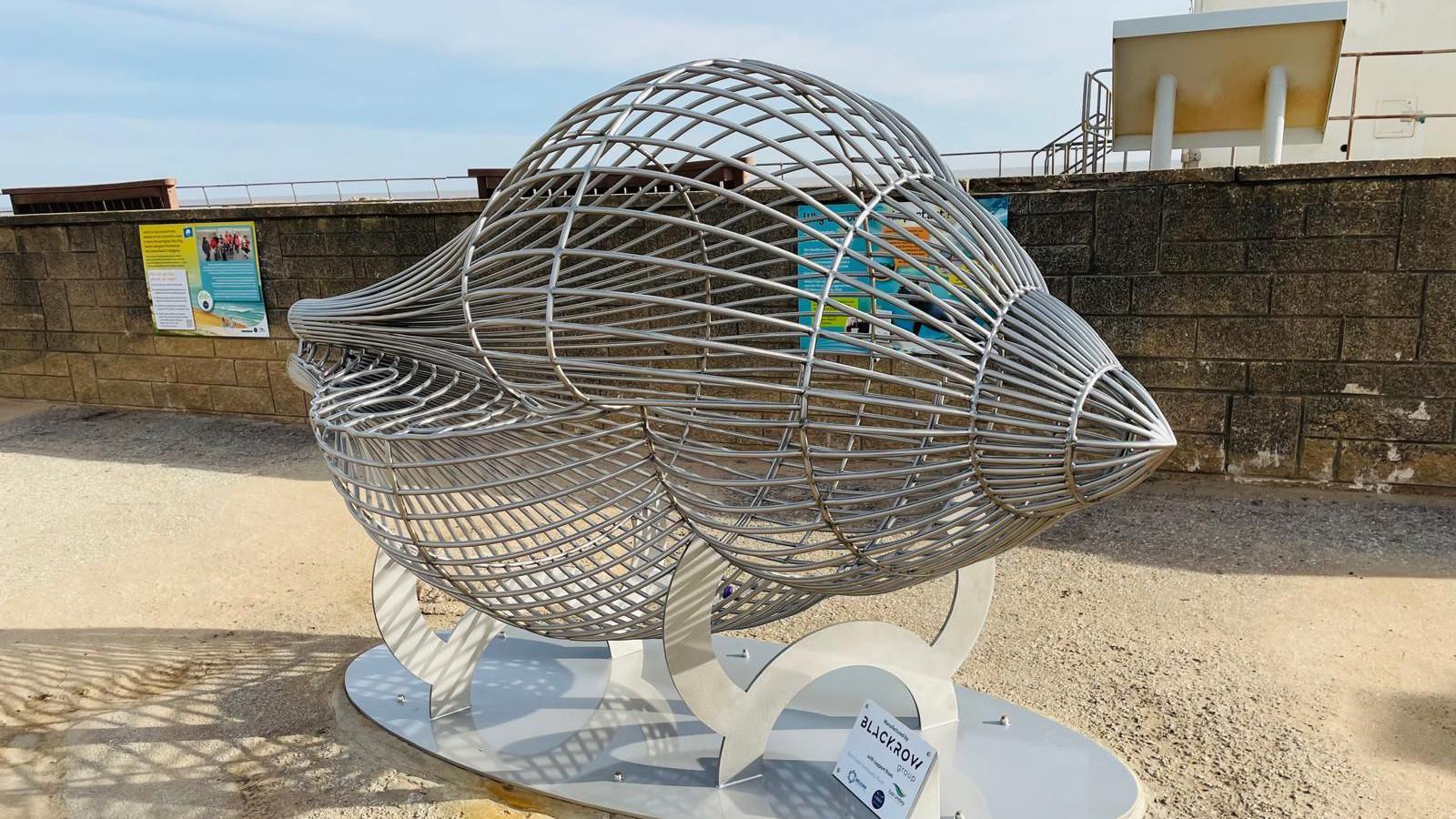 A side view of the large metal seashell recycling bin on the promenade of Sutton on Sea. A plaque is attached at the bottom which reads 'Blackrow Group'.