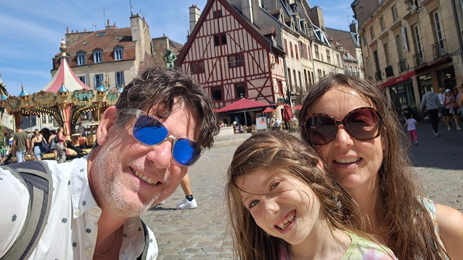 Photo of Aisha's dad, Aisha and Lucy smiling in a family picture. On the left, you can see a fairground carousel in the background. There is a town centre in the background behind the family.