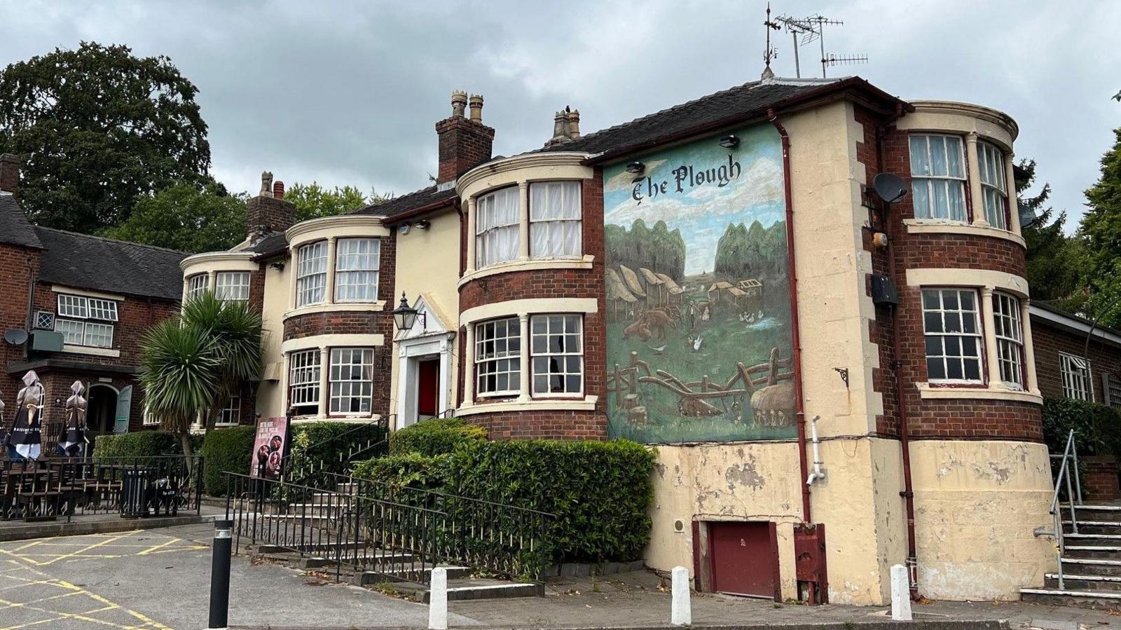 A large mural on the side of a brick-built building. There is cream-coloured rendering around much of the building and a large mural painted on the large flat surface on the right hand side of the building.