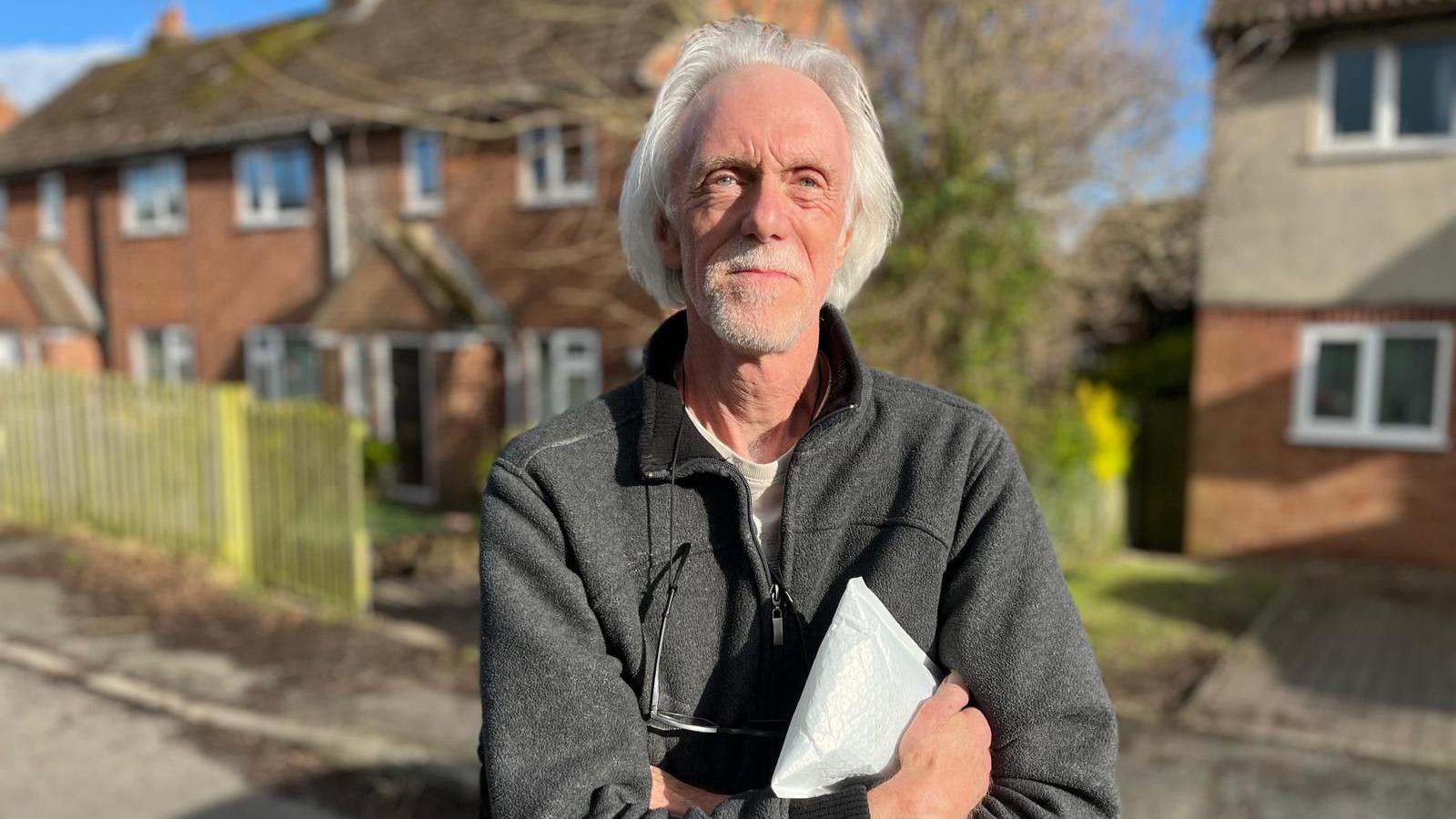 Paul Rowland, with white hair brushed back and wearing a grey fleece, stands smiling at the camera with his arms folded.