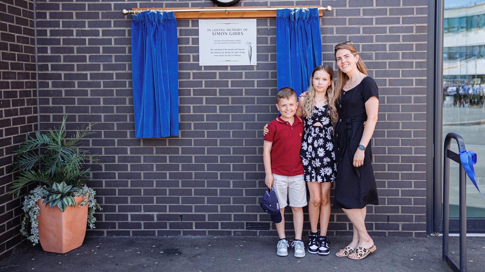 Two children and their mum wearing a black dress standing next to the Simon Gibbs plague