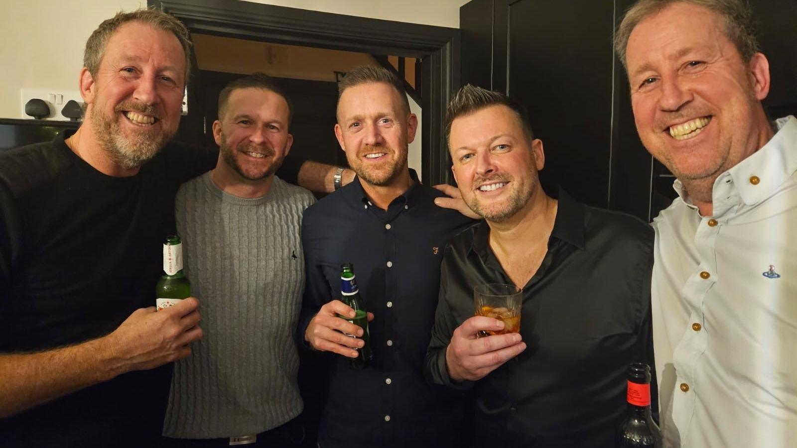 Five men gathered close together, holding drinks with big smiles. They all have a drink in hand and appear to be in a pub.