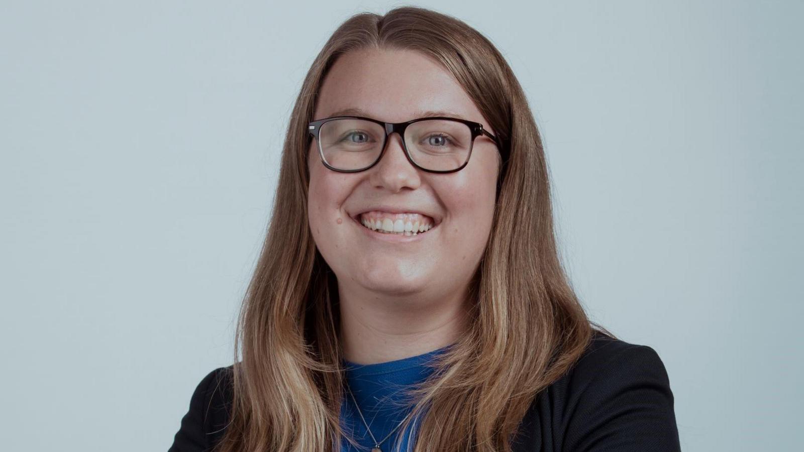 Rachel Hayden is pictured. She has long brown hair and glasses. She is wearing a blue shirt with a black blazer over the top. She is looking at the camera and smiling.
