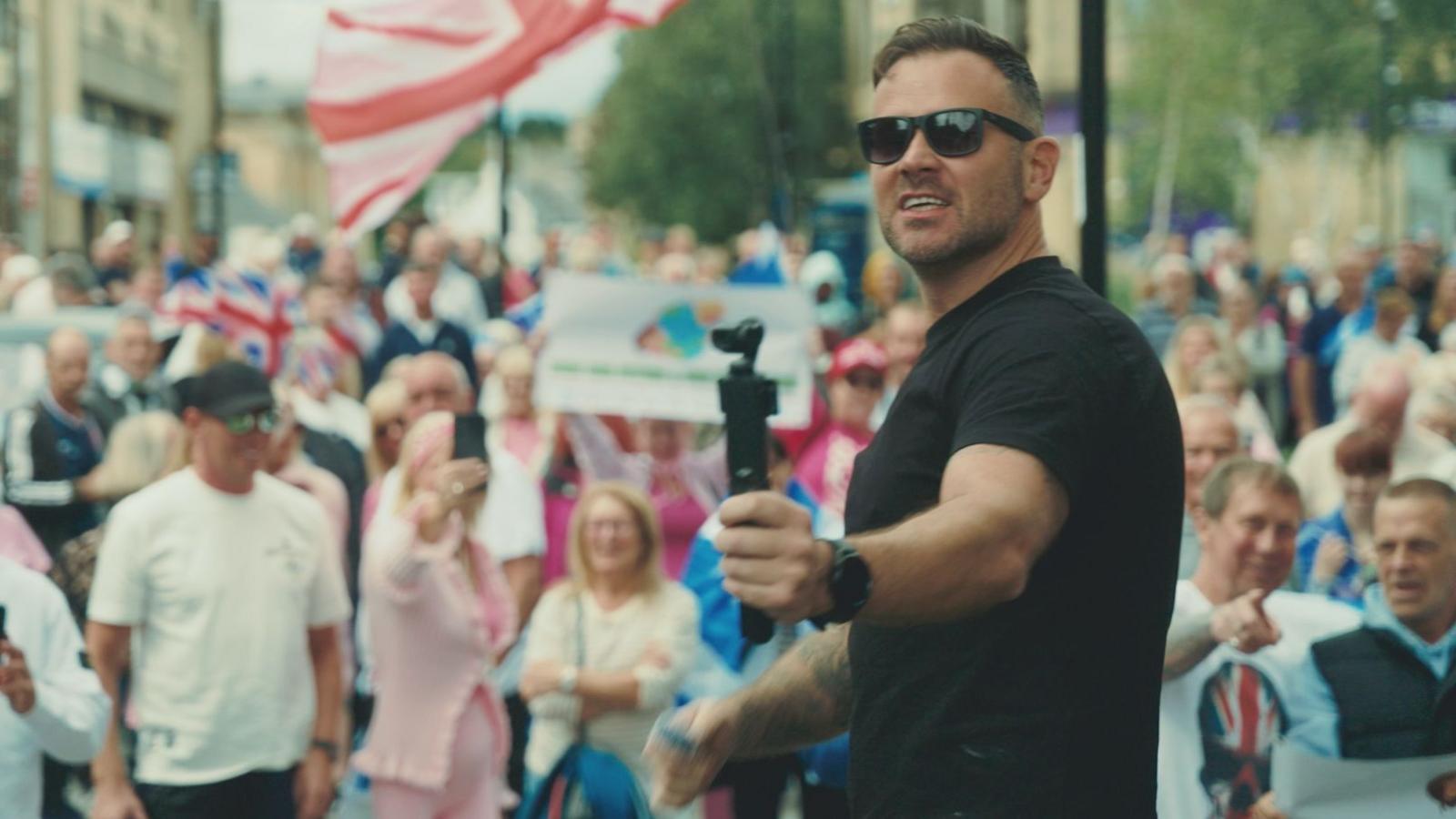 John Watt standing in front of a crowd of people, some of who are holding union flags. He has short brown hair and is wearing a black tshirt and sunglasses, while holding what looks like a selfie stick.