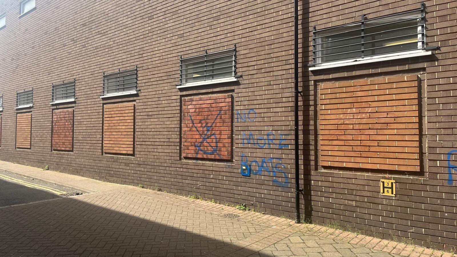 Brown brick building with graffiti on it saying 'no more boats' in blue paint