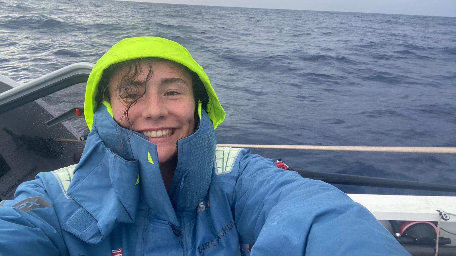 Zara Lachlan wearing a light blue coat with a neon yellow hood while on her boat in the middle of the ocean. She is smiling at the camera and a strand of her hair has come out from under her hood. It has fallen across her freckled face and is wet.
