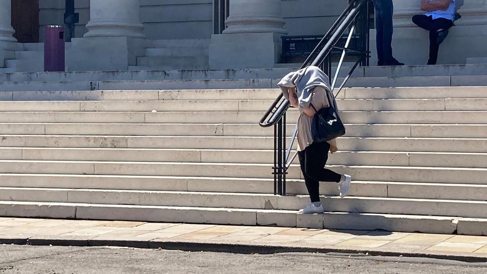 Steps of crown court and woman walking down last steps in black jeans and white trainers with a black shoulder bag and loose beige top. She is holding a grey jumper over head to cover her face and hair.