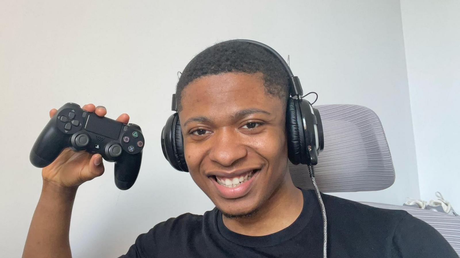 Dienne, a man with short hair, smiles into the camera wearing big gaming headphones. He's wearing a black t-shirt sat on a grey chair with a head rest. He's holding up a black Playstation 2 controller.