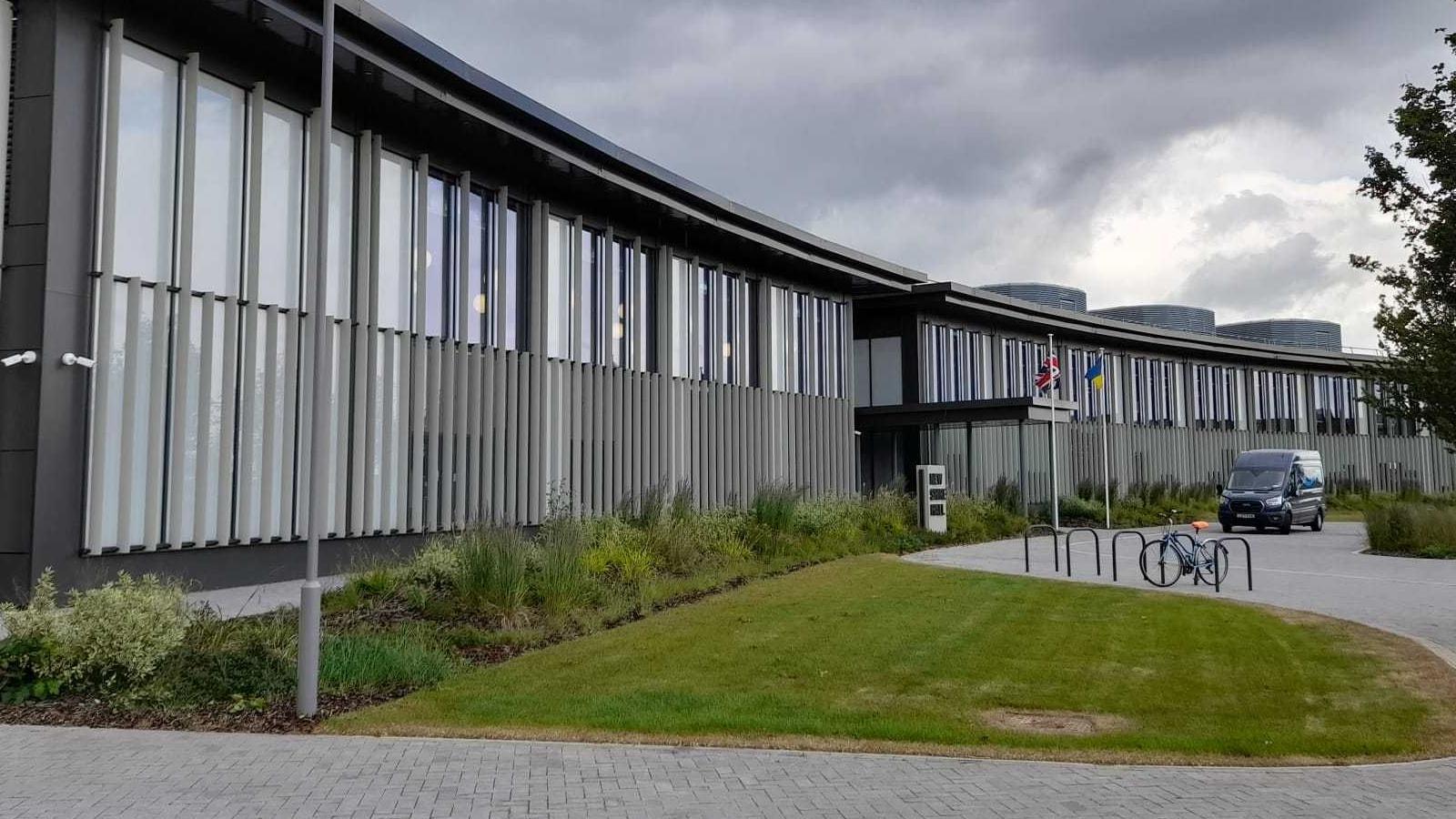 New Shire Hall in Alconbury Weald, Cambridgeshire is a slightly curved crescent shaped building with grey exterior walls and light grey stripes with windows on the front. There are two flags, Great British and Ukrainian, flying at the front. This is the base for Cambridgeshire County Council.