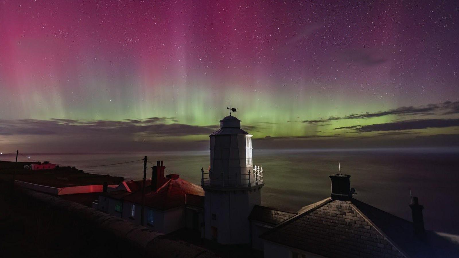 Pink, purple, yellow and green lights in the night sky above the sea with a lighthouse and house roofs in the foreground.