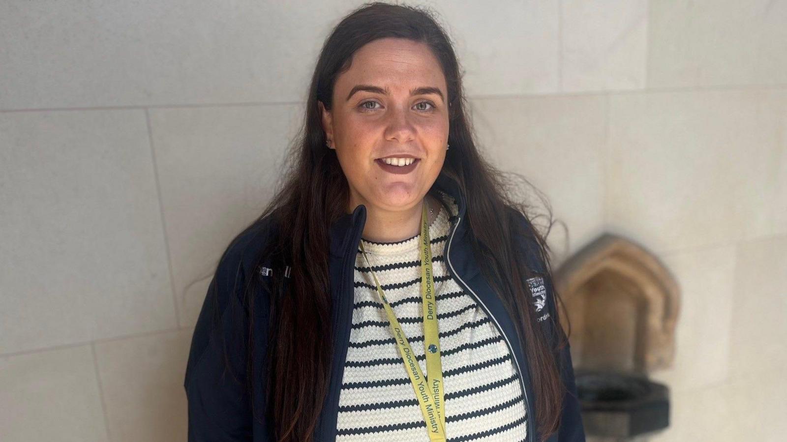 Lizzie Rae is a young woman with long brown hair. She is wearing a navy zip up top over a white and navy striped jumper. She is staring at the camera and smiling.