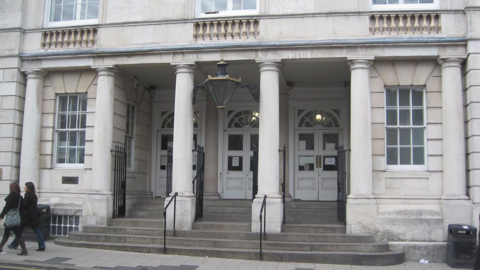 The exterior of Lewes Crown Court. A light grey, stone building with three large white doors. Two people walk right to left on the pavement in front of the building